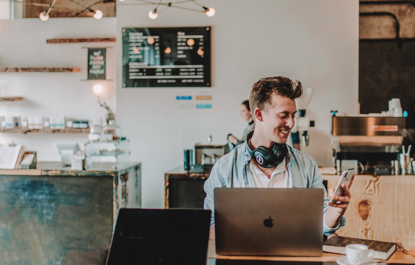 A young business owner smiling at his phone in a cafe, laptop open